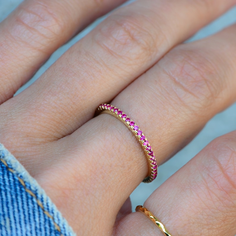 Puede incluir: Un fino anillo de banda de oro con una fila de peque&ntilde;as piedras preciosas rosas.