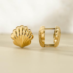 May include: A pair of gold-toned hoop earrings featuring a three-dimensional shell design. One earring is positioned to show the front view of the scalloped shell shape, while the other is oriented to display the hinged closure mechanism.
