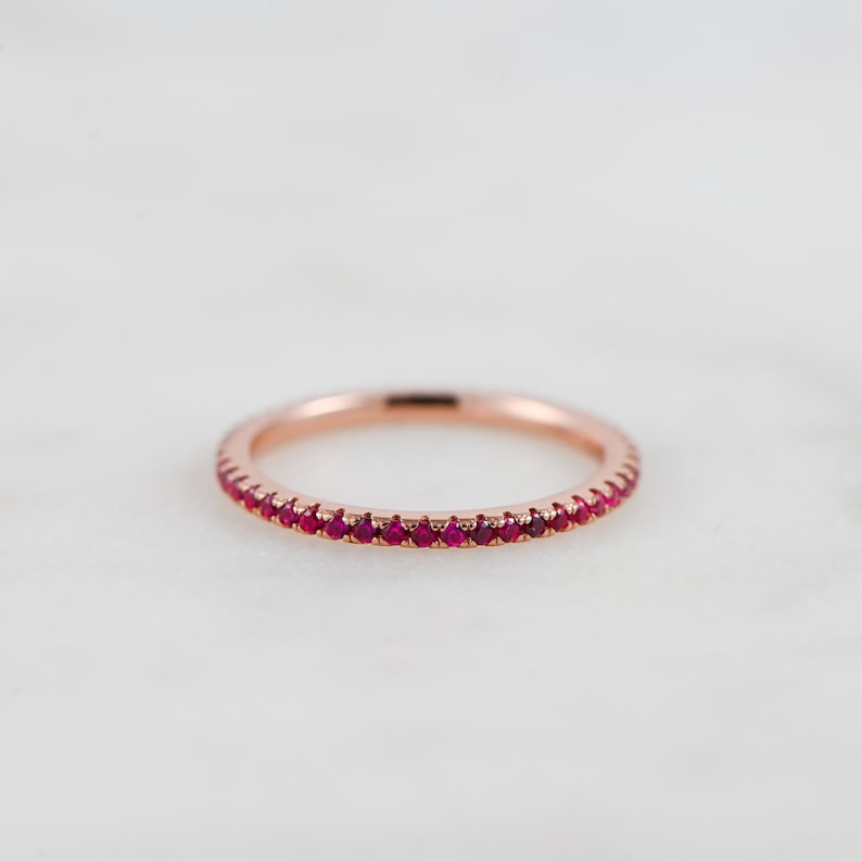 Puede incluir: Un delicado anillo de banda de oro rosa con una fila de peque&ntilde;as piedras de rub&iacute; rojo brillante.