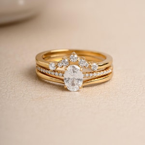 May include: A set of gold rings with diamond accents. The engagement ring features an oval-cut diamond. The set includes a diamond-studded band and a curved ring with a row of diamonds, creating a layered look. The rings are displayed on a neutral surface.