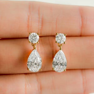 May include: A pair of gold-toned drop earrings. Each earring features a round, clear gemstone at the top and a larger, pear-shaped clear gemstone suspended below. The earrings are held in a person's hand.