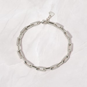 May include: A silver chain necklace with a heart-shaped clasp. The chain is made of linked rectangular pieces, each with a row of small clear stones.