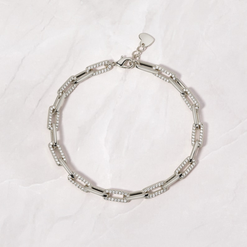 May include: A silver chain necklace with a heart-shaped clasp. The chain is made of linked rectangular pieces, each with a row of small clear stones.