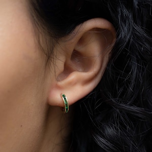 May include: A pair of gold hoop earrings with a row of emerald green gemstones set into the band.