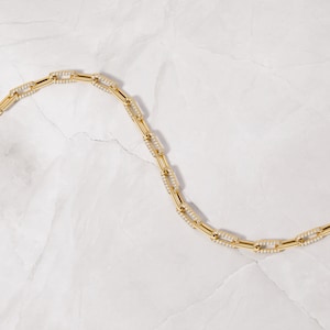 May include: A gold chain necklace with small, square-shaped links that are encrusted with tiny clear stones. The necklace is laying on a white, textured surface.