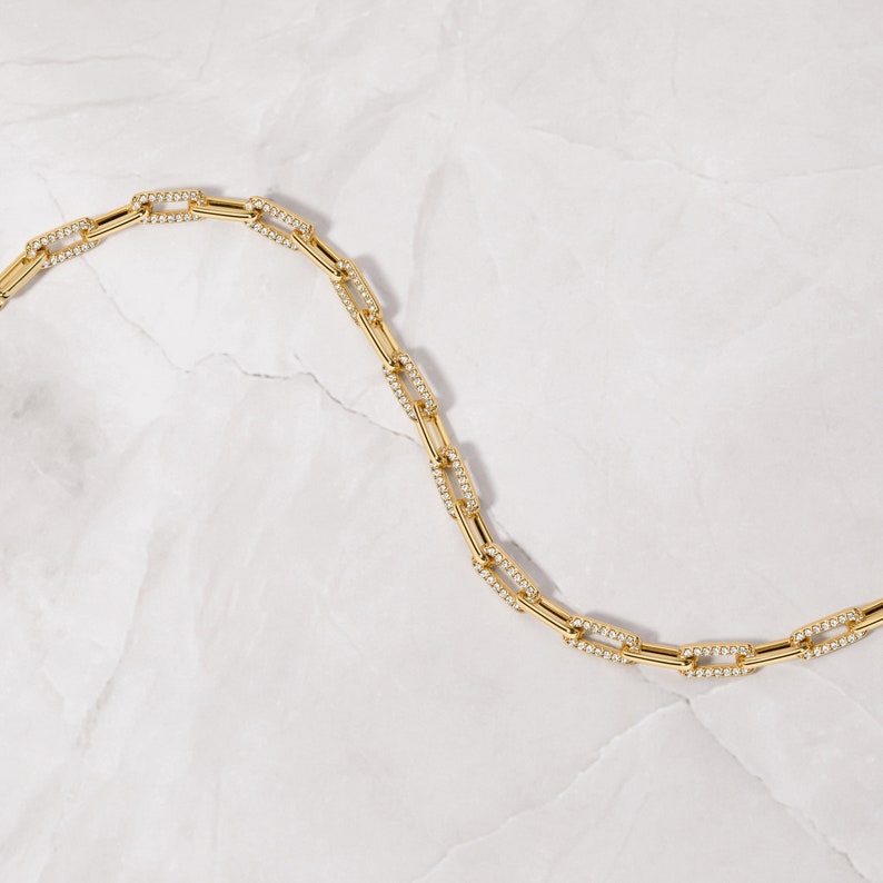 May include: A gold chain necklace with small, square-shaped links that are encrusted with tiny clear stones. The necklace is laying on a white, textured surface.