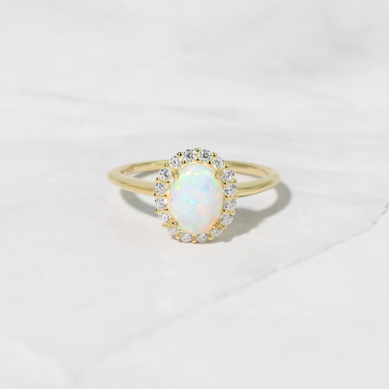 Pode incluir: Um anel de ouro com uma gema de opala branca cercada por um halo de diamantes menores.