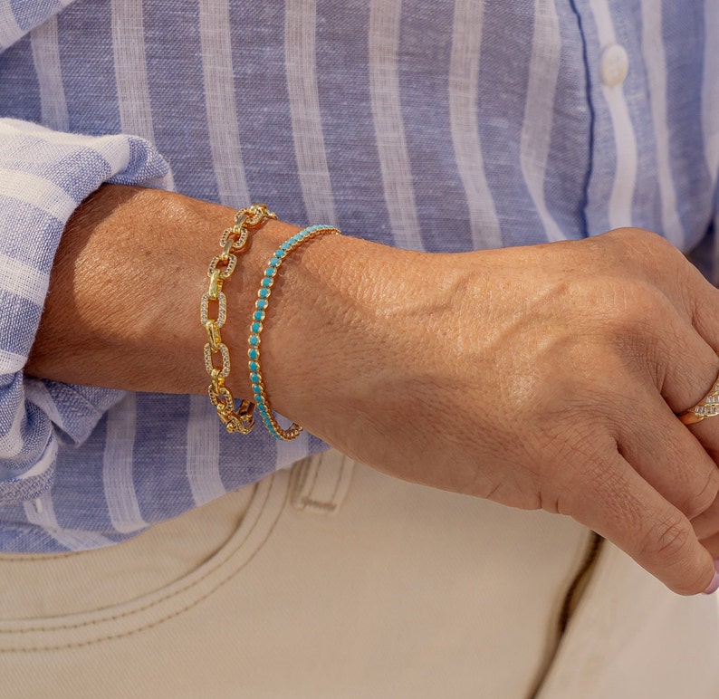 May include: Two gold bracelets on a person's wrist. One bracelet is a chain link style with clear stones, the other is a thin band with turquoise stones.