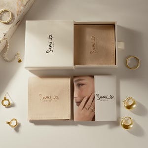 Puede incluir: Vista cenital de joyas de oro, que incluyen anillos, pendientes y una pulsera con un dije de coraz&oacute;n. Las joyas se exhiben con cajas blancas y bolsas beige, todas con el logotipo "Sami Jewels".