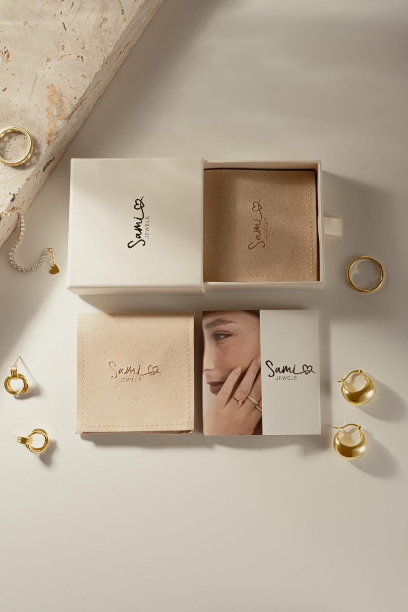 Puede incluir: Vista cenital de joyas de oro, que incluyen anillos, pendientes y una pulsera con un dije de coraz&oacute;n. Las joyas se exhiben con cajas blancas y bolsas beige, todas con el logotipo "Sami Jewels".