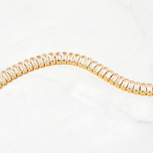 Pu&ograve; includere: Un braccialetto a catena d'oro con cristalli trasparenti a forma di baguette.