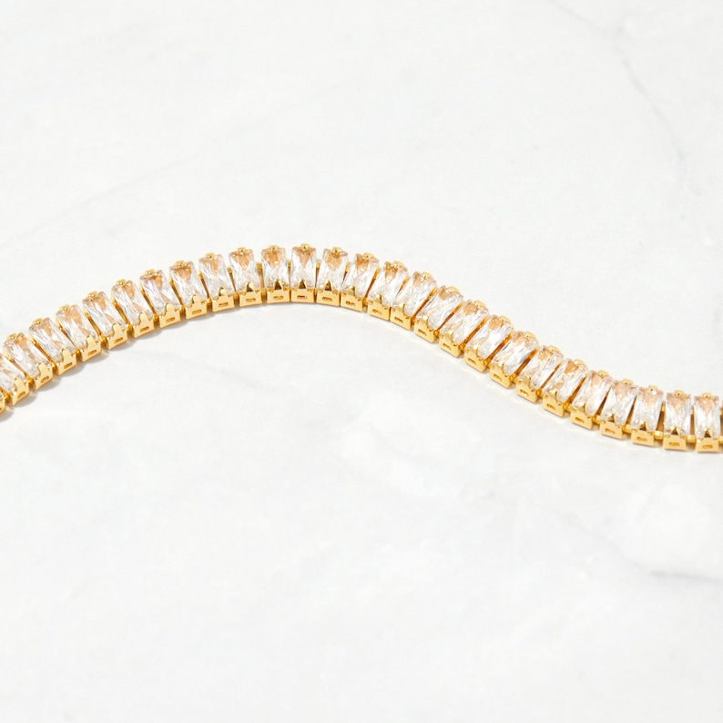 Pu&ograve; includere: Un braccialetto a catena d'oro con cristalli trasparenti a forma di baguette.