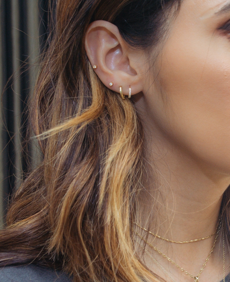 Puede incluir: Una oreja de mujer con tres pendientes de oro. Los pendientes son un peque&ntilde;o pendiente de bot&oacute;n, un pendiente de bot&oacute;n m&aacute;s grande con un peque&ntilde;o diamante y un peque&ntilde;o aro de oro con una fila de peque&ntilde;os diamantes.