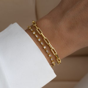 May include: Three gold bracelets with clear cubic zirconia stones. The top bracelet has a chain link design, the middle bracelet has a square stone design, and the bottom bracelet has a rectangular stone design.