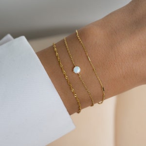 May include: Three gold chain bracelets with a white opal gemstone on one of the chains. The bracelets are layered on a person's wrist.