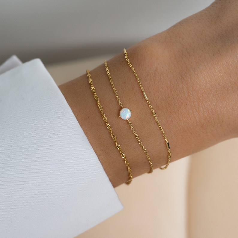 May include: Three gold chain bracelets with a white opal gemstone on one of the chains. The bracelets are layered on a person's wrist.