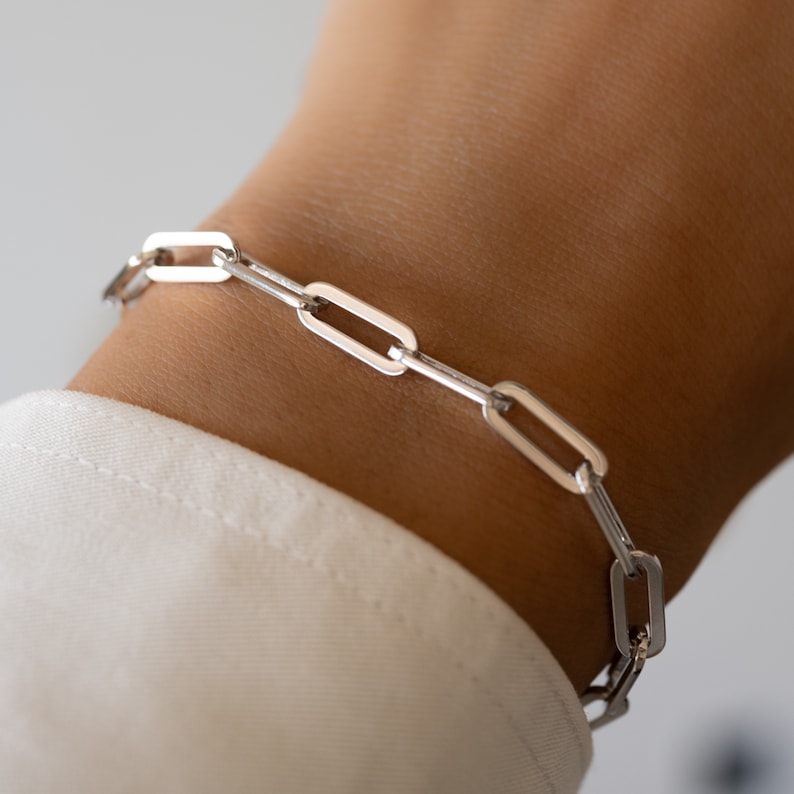 May include: A silver chain bracelet with a link style design. The bracelet is on a person's wrist.