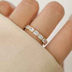 May include: A gold ring with a series of rectangular, clear gemstones set in a row. The ring is on a finger, with a neutral-colored fabric in the background. The ring's design is simple and elegant.