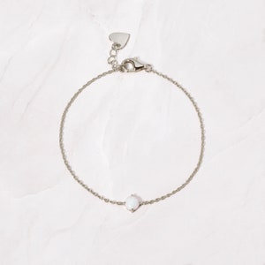 May include: A delicate silver chain bracelet with a small white opal gemstone. The bracelet has a heart-shaped clasp.