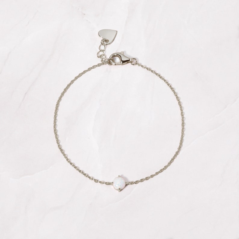 May include: A delicate silver chain bracelet with a small white opal gemstone. The bracelet has a heart-shaped clasp.