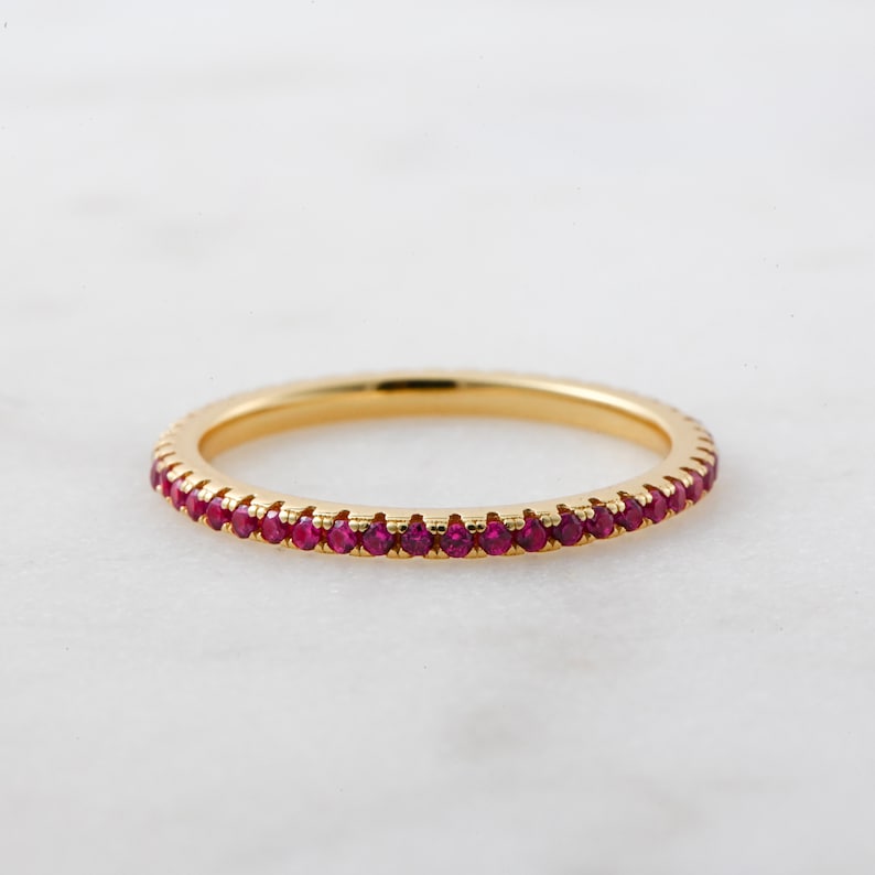 Puede incluir: Un delicado anillo de banda de oro con una fila de peque&ntilde;as piedras de rub&iacute; rojas.