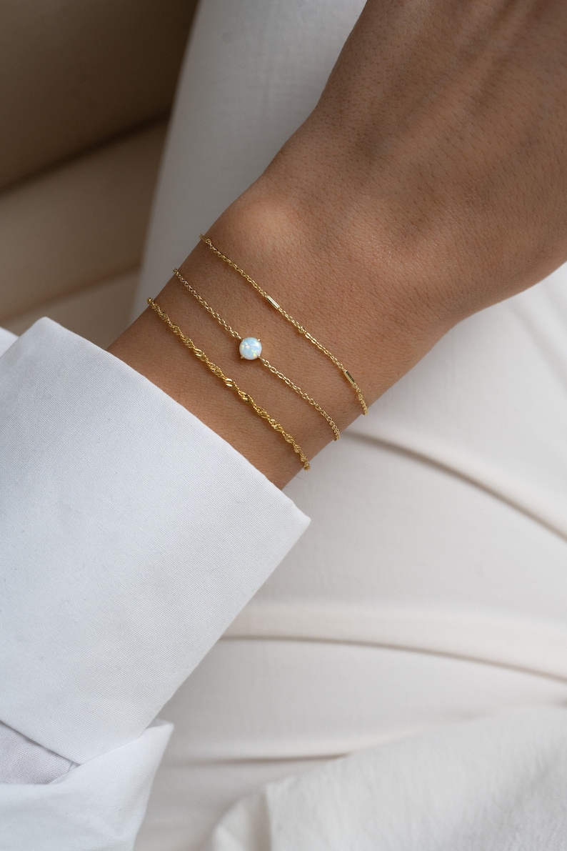 May include: A set of three gold bracelets on a wrist. The top bracelet is a delicate chain with a small opal gemstone. The middle bracelet is a thicker chain with a rope-like design. The bottom bracelet is a thin chain.