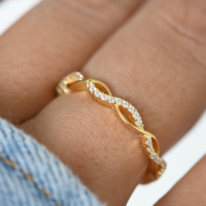 May include: A gold ring with a woven design, set with small, clear gemstones. The ring is worn on a finger, highlighting its intricate details and sparkling appearance. The ring's design features a continuous pattern of interwoven loops.