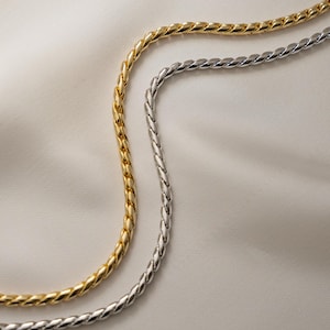 Puede incluir: Primer plano de dos collares de cadena trenzada. Uno es de color dorado, el otro plateado. Las cadenas están sobre una superficie blanca ligeramente texturizada. Las cadenas tienen un aspecto trenzado.