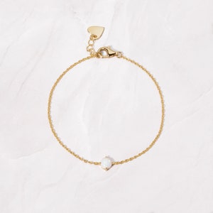 May include: A delicate gold chain bracelet with a small white opal gemstone. The bracelet has a heart-shaped clasp.