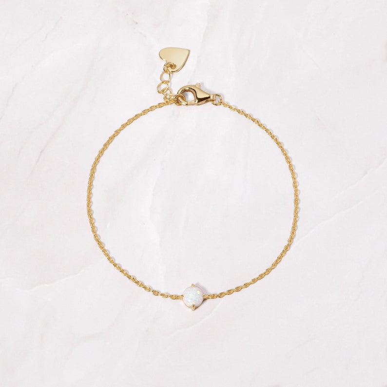 May include: A delicate gold chain bracelet with a small white opal gemstone. The bracelet has a heart-shaped clasp.