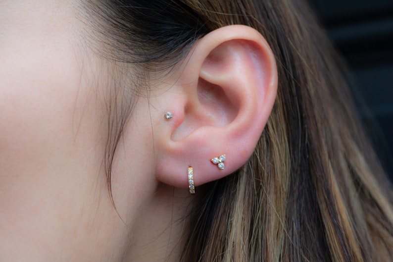 May include: A person's ear with three different earrings. The top earring is a small stud, the middle earring is a small cluster of three stones, and the bottom earring is a small hoop with stones.