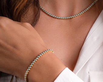 May include: Gold-toned necklace and bracelet set. Both pieces feature small, round, light blue gemstones. The jewelry is displayed against a white shirt.