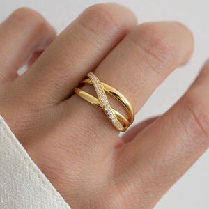 Op de afbeelding: Een goudkleurige ring met een verweven ontwerp. Eén band is bezet met kleine, heldere edelstenen. De ring zit om een vinger, met witte stof op de achtergrond. De ring is een sieraad.