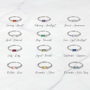 May include: Twelve silver rings with different colored gemstones, each representing a different month of the year. The rings are arranged in three rows of four, with the month and gemstone name written below each ring.