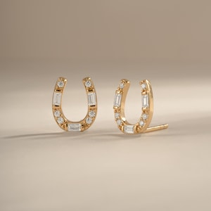 May include: A pair of gold horseshoe-shaped stud earrings. Each earring features a series of small, round, clear stones set along the curve, accented by three rectangular baguette-cut clear stones positioned at the base and sides of the horseshoe.