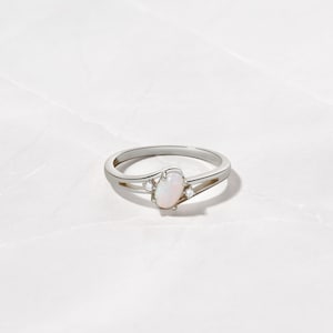 May include: A silver ring with an oval opal center stone and two small accent stones. The ring has a split shank design and is set against a white background. The opal has a soft, milky appearance.