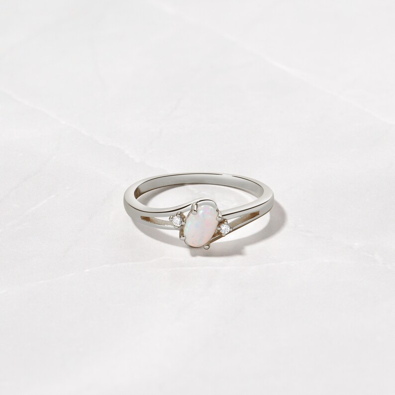 May include: A silver ring with an oval opal center stone and two small accent stones. The ring has a split shank design and is set against a white background. The opal has a soft, milky appearance.