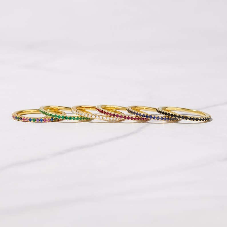 May include: Six gold rings with a single row of colorful gemstones. The rings are arranged in a row, with the black gemstone ring on the far right and the green gemstone ring on the far left.