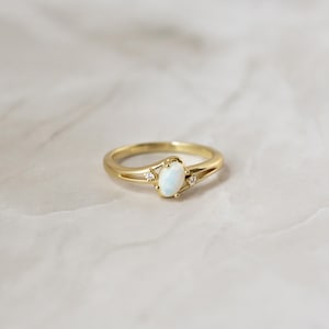 May include: A gold ring with an oval, opalescent center stone and two small accent stones. The band has a split design on either side of the center stone. The ring is set against a neutral background.