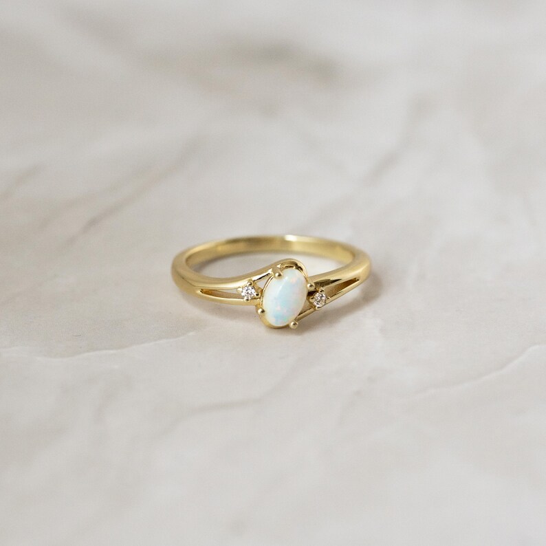 May include: A gold ring with an oval, opalescent center stone and two small accent stones. The band has a split design on either side of the center stone. The ring is set against a neutral background.