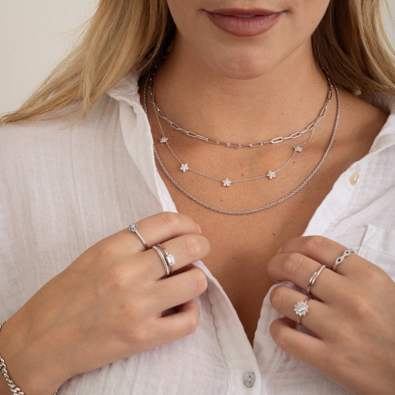 Pu&ograve; includere: Una donna indossa una camicia bianca con una collana a catena d'argento e una catena d'argento con piccoli ciondoli a forma di fiore. Indossa diversi anelli d'argento alle dita.