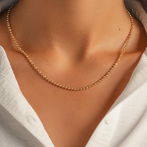 May include: A delicate gold necklace with a beaded chain. The necklace is worn against a white collared shirt. The chain is made of small, round beads that catch the light, creating a subtle sparkle. The necklace is a simple yet elegant piece of jewelry.