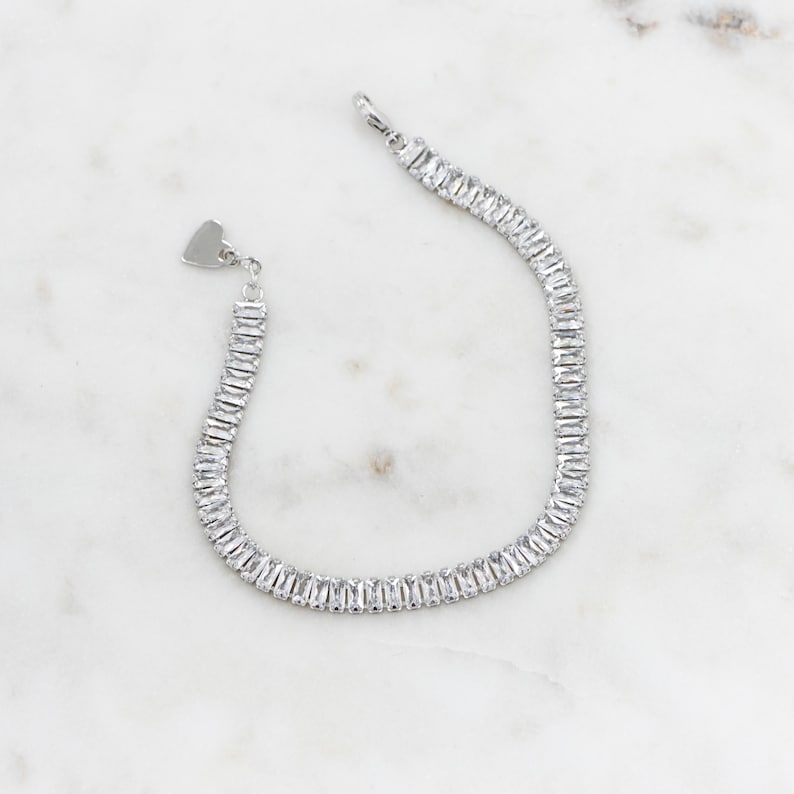 Pu&ograve; includere: Un braccialetto d'argento con un ciondolo a forma di cuore e una catena di pietre chiare a forma di baguette.