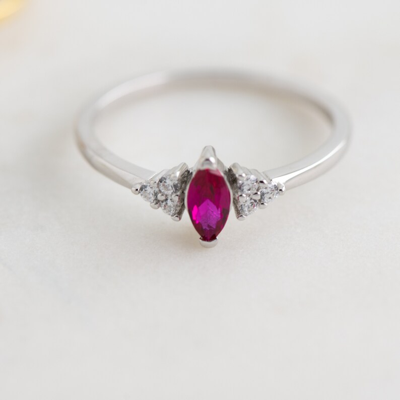 K&ouml;nnte beinhalten: Ein silberner Ring mit einem rubinroten Marquise-Schliff und kleineren, klaren Edelsteinen auf der Ringschiene.