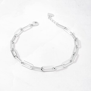 May include: A silver chain bracelet with a heart-shaped clasp. The chain is made of interlocking rectangular links.