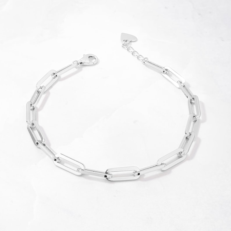 May include: A silver chain bracelet with a heart-shaped clasp. The chain is made of interlocking rectangular links.