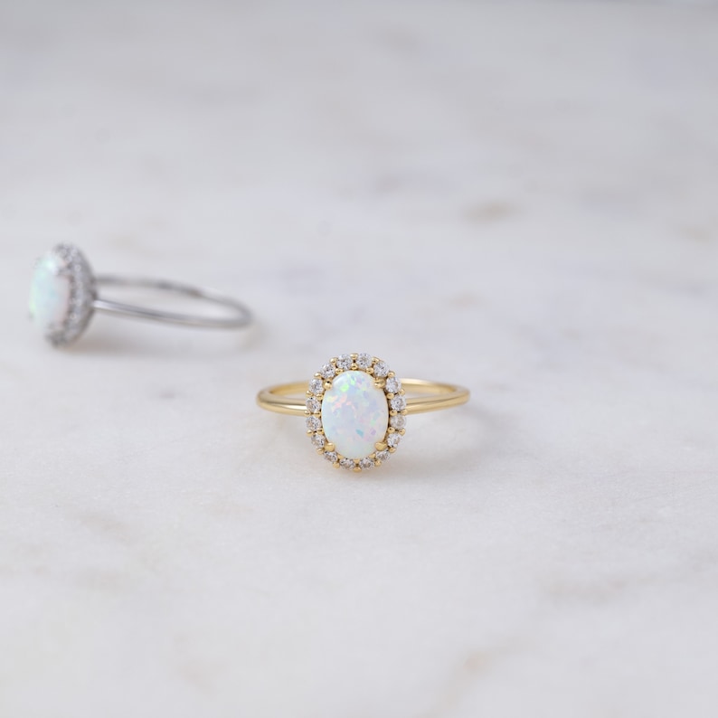 Pode incluir: Um anel de ouro com uma pedra de opala branca cercada por um halo de pedras preciosas menores e transparentes.