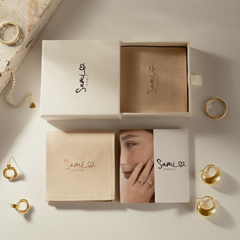 May include: A white box with a gold logo that says "Sami Jewels" on the front. The box is open and there is a smaller white box inside with a beige velvet lining. The smaller box also has the "Sami Jewels" logo on the front. There are several gold jewelry pieces on a white surface around the boxes.