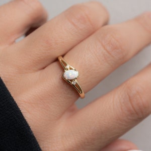 May include: A gold ring featuring an oval opal center stone and two small accent diamonds. The ring has a delicate, split-shank design. The opal has a milky white appearance. The ring is a piece of jewelry.