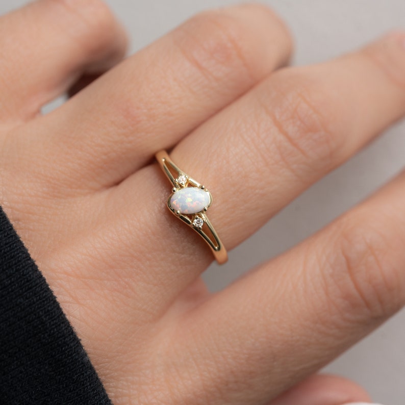 May include: A gold ring featuring an oval opal center stone and two small accent diamonds. The ring has a delicate, split-shank design. The opal has a milky white appearance. The ring is a piece of jewelry.